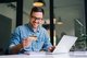 Smiling man at a desk in front of a laptop computer holding a payment card. [Image by creator Moon Safari from Adobe Stock]